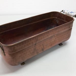 May include: A copper planter with a hammered finish and two white ceramic handles with blue floral designs. The planter has four small feet and is rectangular in shape.
