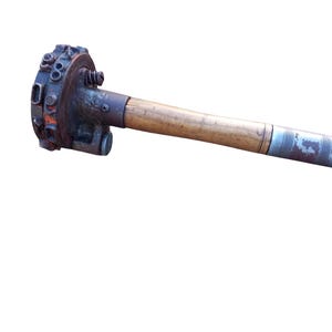 Puede incluir: Un componente de máquina de escribir vintage tipo rueda de margarita con una cabeza metálica circular con caracteres en relieve, unido a un mango largo de madera con una pieza final de metal plateado texturizado, mostrando signos de edad y oxidación.