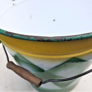 Puede incluir: Un cubo de esmalte vintage con un dise&ntilde;o amarillo, blanco y verde. El cubo tiene un asa de madera y un borde de alambre met&aacute;lico.
