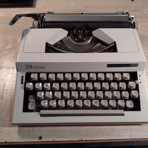 Pode incluir: Uma máquina de escrever vintage branca com um teclado preto. A máquina de escrever tem o nome da marca "TA ROYAL" e o nome do modelo "contassa 2" impresso nela.