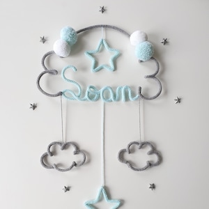 May include: A gray and light blue knitted mobile with the name "Sean" spelled out in light blue yarn. The mobile features a cloud shape with two pom-poms, a star, and two hanging clouds and a star.