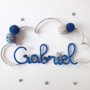 May include: A blue and grey yarn name banner with the name "Gabriel" spelled out in blue yarn. The banner is shaped like a cloud and has three pom-poms attached to it.