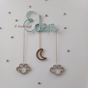 May include: A light blue crocheted name sign that says "Eden" with a brown crescent moon and two beige clouds hanging from it. The sign is decorated with silver glitter stars.