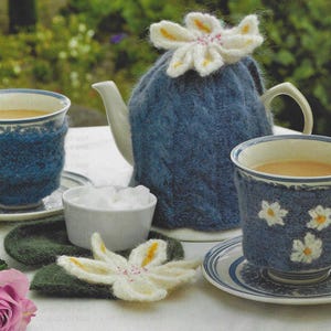 May include: A blue knitted teapot cosy with a white crocheted flower on top. Two blue teacups with white trim and knitted cosies are on a table with a white saucer and a bowl of sugar cubes.