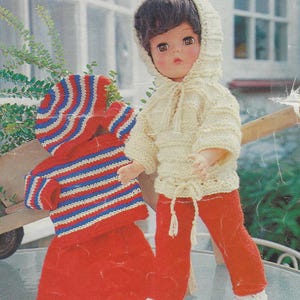 May include: A doll wearing a knitted outfit. The outfit includes a white hooded sweater, red trousers, and a striped red, white and blue sweater with a matching hat.