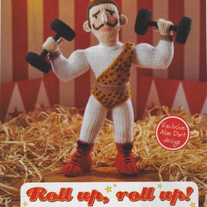 May include: A knitted strongman doll, wearing a white top, brown trunks, and red boots, lifting black dumbbells. The doll has a mustache and is set against a circus backdrop. Text reads "Roll up, roll up!" and "Exclusive Alan Dart design."