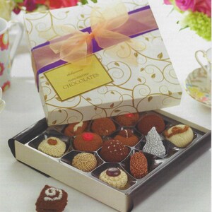May include: A square gift box with a white and gold swirl pattern, a purple ribbon, and a peach bow. The label reads "delicious HAND-KNITTED CHOCOLATES". Inside are various round, textured, knitted items resembling chocolates, with one piece placed outside.