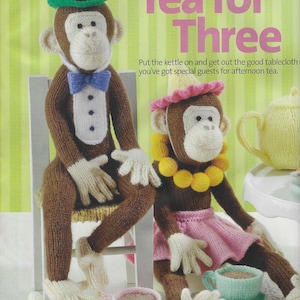 May include: Two knitted monkey dolls sitting at a table with tea sets. One monkey wears a green top hat and blue bow tie. The other wears a pink hat, yellow necklace, and pink skirt. A yellow knitted banana sits nearby. Text reads Tea for Three.
