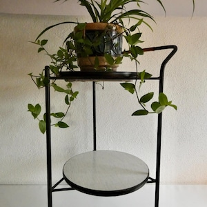 Könnte beinhalten: Ein schwarzer Pflanzenständer aus Metall mit zwei runden Ablagen und Rollen. Auf der oberen Ablage steht eine Topfpflanze mit grünem Laub. Die untere Ablage hat ein weißes und graues Muster. Die Ranken der Pflanze ranken herab.