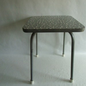 Könnte beinhalten: Kleiner, vintage Beistelltisch mit rechteckiger Tischplatte mit einem Mosaikfliesenmuster in Grau- und Weißtönen. Der Tisch hat gebogene, graue Metallbeine mit weißen Kunststofffüßen. Ein klassisches Möbelstück.
