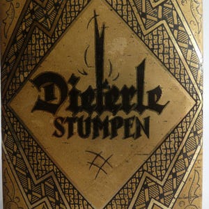 Könnte beinhalten: Nahaufnahme einer rechteckigen Vintage-Box mit goldenem und schwarzem Design. Die Box hat ein rautenförmiges Emblem mit den Worten "Dieterle STUMPEN" in schwarzer gotischer Schrift. Aufwendige geometrische Muster umranden das Emblem und die Box.
