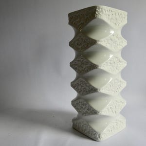 Könnte beinhalten: Eine hohe, cremeweiße Keramikvase mit einem sich wiederholenden geometrischen Muster aus glatten, konkaven Vertiefungen, die sich mit strukturierten, rauen, rautenförmigen Vorsprüngen abwechseln, welche vertikal am Objekt verlaufen.