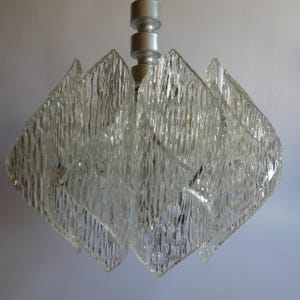 Könnte beinhalten: Ein Kronleuchter aus klarem Glas mit strukturierter Oberfläche. Der Kronleuchter hat einen silberfarbenen Metallstiel und besteht aus mehreren rautenförmigen Glasplatten. Die Platten haben ein strukturiertes, geriffeltes Aussehen.