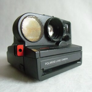 Könnte beinhalten: Eine schwarze Polaroid Land Camera mit einem prominenten Objektiv und einem Lautsprecher-ähnlichen Element auf der Oberseite. Die Kamera hat einen roten Knopf an der Seite und die Worte "POLAROID LAND CAMERA" sind auf der Vorderseite aufgedruckt.