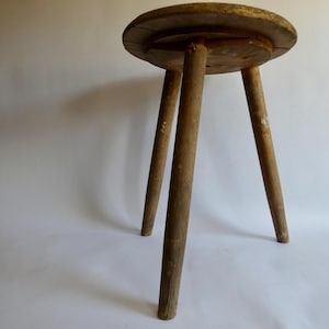 Könnte beinhalten: Ein rustikaler, dreibeiniger Holzschemel mit rundem Sitz. Der Schemel ist aus hellbraunem Holz gefertigt und weist Gebrauchsspuren auf, mit sichtbarer Maserung und einem leicht verwitterten Aussehen. Die Beine sind stabil und nach außen abgewinkelt.