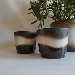 Könnte beinhalten: Zwei Keramik-Pflanztöpfe mit einem braunen, weißen und dunkelbraunen Farbschema. Die Töpfe haben ein horizontales Streifendesign. Ein größerer Topf und eine Pflanze befinden sich im Hintergrund.