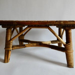 Könnte beinhalten: Ein kleiner, niedriger rechteckiger Hocker aus naturfarbenem, hellbraunem Rattan. Das Möbelstück verfügt über eine geflochtene, flache Sitzfläche, die von vier stabilen Beinen mit Kreuzverstrebungen und festen Rattanwicklungen gestützt wird.