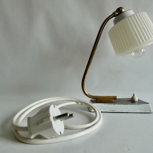 Könnte beinhalten: Eine Schreibtischlampe mit einem geschwungenen Messingarm, einem weißen, gerippten Glasschirm und einem weißen Stromkabel mit zweipoligem Stecker. Der Sockel ist weiß mit einer strukturierten Metalloberfläche und einem kleinen weißen Druckschalter.