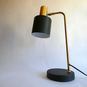 Könnte beinhalten: Eine Schreibtischlampe mit einem mattschwarzen zylindrischen Schirm und einem runden Sockel, verbunden durch einen schlanken, polierten goldfarbenen Metallarm. Die Lampe verfügt über ein zylindrisches goldenes Element am Schirm und ein schwarzes Kabel.