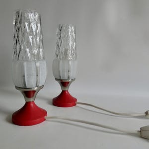 Könnte beinhalten: Zwei Vintage-Tischlampen mit roten Sockeln, silbernen Stielen und transparenten, strukturierten Glasschirmen. Die Lampen haben weiße, gerippte Mittelstücke und sind mit einem weißen Kabel mit Ein-/Ausschalter verbunden. Dekorative Beleuchtung.