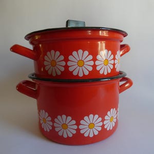 Könnte beinhalten: Zweiteiliges Kochtopf-Set aus rotem Emaille. Die Töpfe sind mit weißen Gänseblümchen mit gelben Zentren verziert. Das Set enthält einen Deckel und hat schwarze Verzierungen und Griffe. Eine charmante Ergänzung für jede Küche.
