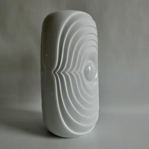 Könnte beinhalten: Eine weiße Keramikvase mit einem einzigartigen Design. Die Vase hat eine zylindrische Form mit einer Reihe konzentrischer, gebogener Rippen, die eine strukturierte Oberfläche erzeugen. Die Rippen konvergieren zur Mitte und bilden ein augenähnliches Muster.