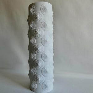 Könnte beinhalten: Weiße Keramikvase mit einem erhabenen Blumenmuster. Die Vase ist hoch und zylindrisch.