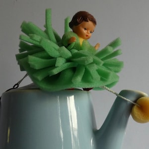 Könnte beinhalten: Eine kleine Puppe mit braunen Haaren und gelbem Oberteil sitzt auf einer hellblauen Teekanne. Die Puppe ist von einem großen, kreisförmigen hellgrünen Schaumstoff-Fransenrock umgeben. Eine dünne weiße Schnur verbindet die Puppe mit einem gelben Schaumstoffkreis am Ausguss.