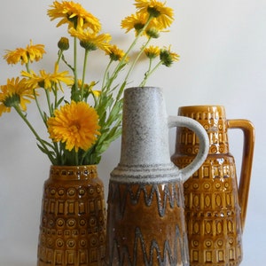 Könnte beinhalten: Drei Keramikvasen in Braun- und Weißtönen mit gelben Blumen. Die Vasen haben geometrische Muster und eine strukturierte Oberfläche. Eine Vase hat einen Griff. Die Blumen sind in einer Vase mit geometrischem Muster.