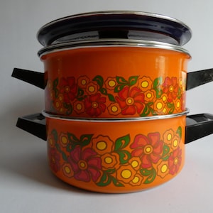 Könnte beinhalten: Zwei orangefarbene Emaille-Töpfe mit schwarzen Griffen und einem dunkelblauen Deckel. Die Töpfe sind mit einem Blumenmuster in Rot, Orange, Gelb und Grün verziert. Die Töpfe sind übereinander gestapelt.