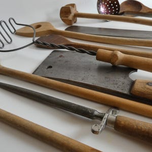 Könnte beinhalten: Eine Sammlung antiker Küchenutensilien, darunter Holzlöffel, ein Fleischbeil, ein Wetzstahl und ein Schneebesen. Die Werkzeuge sind auf einer weißen Oberfläche angeordnet und zeigen ihre natürlichen Holz- und Metallakzente.