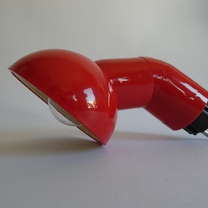 Könnte beinhalten: Eine rote Schreibtischlampe im Retro-Stil mit einem gebogenen Schirm und einem schwenkbaren Arm. Die Lampe hat eine glänzende Oberfläche und eine transparente Glühbirnenabdeckung. Der Stecker hat zwei Stifte. Die Lampe ist für einen Schreibtisch oder Tisch konzipiert.