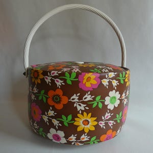 Könnte beinhalten: Ein zylindrischer Aufbewahrungsbehälter mit brauner Basis und einem lebendigen Blumenmuster in Orange, Gelb, Pink und Weiß mit grünen Blättern. Der Behälter hat einen weißen Deckel und einen passenden weißen, bogenförmigen Tragegriff.