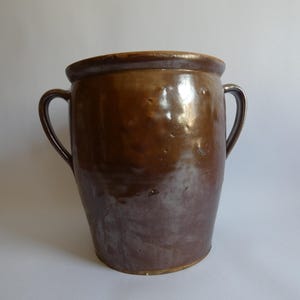 Könnte beinhalten: Ein großer, antiker brauner Steingutkrug mit zwei gebogenen Griffen. Die glasierte Oberfläche hat eine glänzende Oberfläche mit Farbvariationen, die von hellbraun bis dunkelbraun reichen. Der Krug hat eine weite Öffnung und einen abgerundeten Rand.