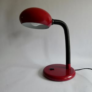 Könnte beinhalten: Eine rote Schreibtischlampe mit einem abgerundeten, ovalen Kopf und einem flexiblen schwarzen Hals. Die Lampe hat einen runden roten Sockel mit einem schwarzen Schalter auf der Oberseite. Das Innere des Lampenkopfes ist weiß und ein schwarzes Kabel führt heraus.