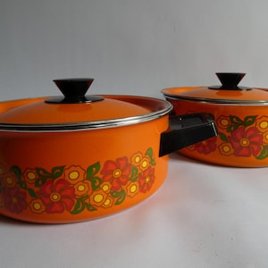 Könnte beinhalten: Zwei leuchtend orangefarbene Emaille-Kochtöpfe mit passenden Deckeln, verziert mit einem Blumenmuster aus roten, gelben und grünen Blüten am Boden. Jeder Topf hat zwei seitliche Griffe aus schwarzem Kunststoff und einen schwarzen Deckelknopf.