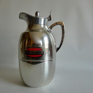 Könnte beinhalten: Eine glänzende, silberfarbene Vintage-Thermoskanne mit Griff und Deckel. Die Thermoskanne hat ein Etikett mit rotem und schwarzem Text "Qualitic". Sie ist zum Warm- oder Kalthalten von Flüssigkeiten bestimmt.