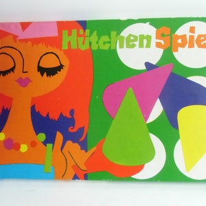 Könnte beinhalten: Eine Vintage-Spieleschachtel mit grünem Hintergrund und dem Text "Hütchen Spiel". Die Schachtel zeigt eine bunte Illustration einer Frau mit orangefarbenen Haaren und einem blauen Kleid.