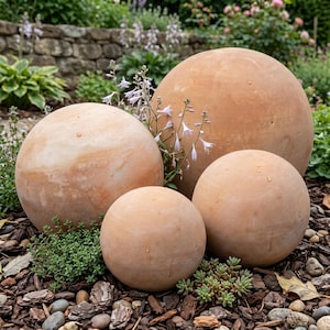 May include: Four spherical, terracotta-colored garden spheres of varying sizes are arranged outdoors. The spheres are smooth and have a natural, earthy appearance. They are surrounded by greenery, including small plants and mulch, creating a natural garden setting.