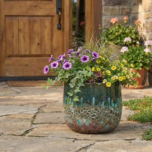 May include: A large, round ceramic planter filled with vibrant flowers. The planter has a teal and brown glazed finish with a textured pattern. Purple, yellow, and green plants spill over the edges, creating a colorful display. The planter sits on a stone patio.