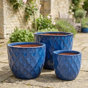 Puede incluir: Tres macetas de cerámica azules con un patrón de diamantes texturizado. Las macetas son de diferentes tamaños y tienen un interior de color terracota. Están colocadas en un patio de piedra con un fondo de jardín.
