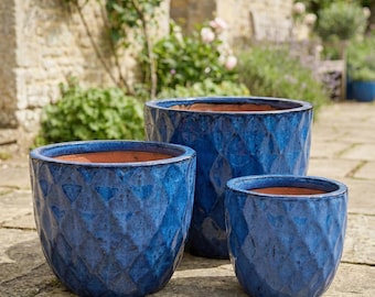 May include: Three blue ceramic planters with a textured diamond pattern. The planters are different sizes and have a terracotta-colored interior. They are set on a stone patio with a garden background.