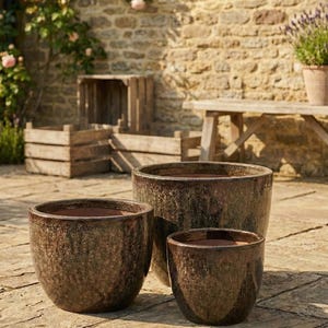 Puede incluir: Tres macetas de cerámica marrón de diferentes tamaños. Las macetas tienen un acabado texturizado y moteado, con un interior marrón oscuro. Se colocan al aire libre en un patio de piedra, con una pared de ladrillo y estructuras de madera en el fondo.