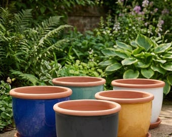 May include: A collection of five ceramic plant pots in blue, dark gray, yellow, teal, and white, each featuring a natural terracotta rim and a matching saucer. The text Exklusive Keramik-Sets mit passenden Untertellern is visible at the top.