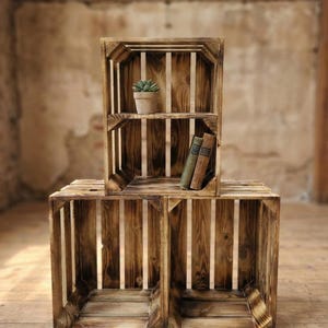 Op de afbeelding: Drie rustieke houten kisten, gerangschikt om een gelaagde presentatie te creëren. Eén kist bevat een kleine potplant en twee vintage boeken. De kisten zijn gemaakt van verweerd hout met verticale lamellen.