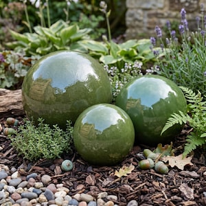 May include: Three glossy, olive-green ceramic spheres of varying sizes are arranged in a garden setting. The spheres reflect the surrounding foliage and sky. The spheres are set among mulch, small stones, and greenery, creating a decorative garden accent.