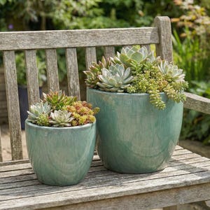 Könnte beinhalten: Zwei türkisfarbene Keramik-Pflanzgefäße mit Sukkulenten auf einer Holzbank. Die Töpfe haben eine glänzende Oberfläche und unterschiedliche Größen. Die Sukkulenten sind eine Mischung aus Grün, Rot und Gelb.