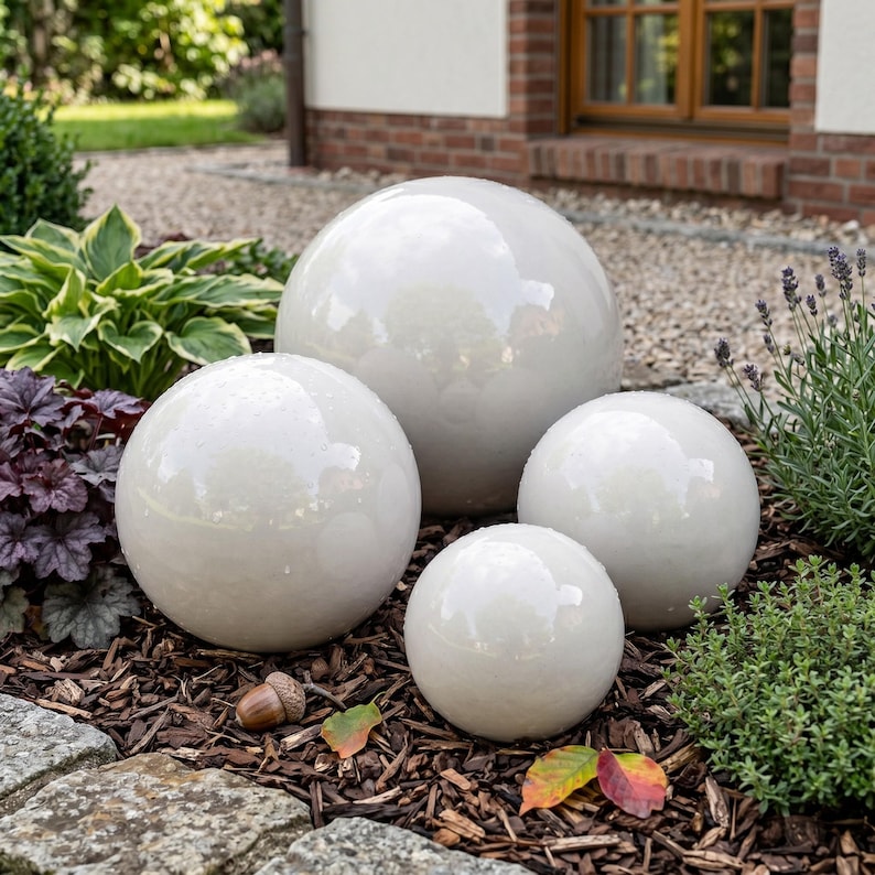 May include: Four white ceramic spheres of varying sizes are arranged on a bed of wood chips. The spheres have a glossy finish and reflect the surrounding greenery. The spheres are a decorative garden accent.