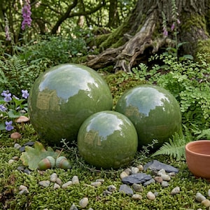 May include: Three glossy, green ceramic spheres of varying sizes are arranged on a bed of moss, surrounded by greenery and small stones. The spheres reflect the surrounding environment, creating a natural, garden-themed aesthetic.