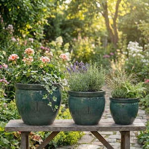 Puede incluir: Tres macetas de cerámica vidriada de color verde azulado de diferentes tamaños se asientan sobre un banco de madera desgastada. Las macetas contienen plantas con follaje verde y flores rosas. El fondo es un jardín exuberante con un sendero de piedra.